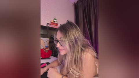 Video of a topless woman with long blonde hair, glasses, lying on a bed, reading a book. Background features a colorful stack of books, a red handbag, and dark curtains.