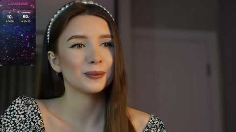 Media: Video of a young woman with fair skin, long brown hair, wearing a black and white floral dress, and a sparkling headband. Background shows a dimly lit room with a digital clock displaying 10:06.