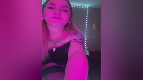 Media: Video of a young woman with long blonde hair, wearing a black top, standing in a dimly lit room with pink and purple LED lights, taking a selfie.