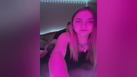 Media: A video of a young woman with long, blonde hair, wearing a black top, taking a selfie in a dimly lit bedroom with pink and purple lighting.