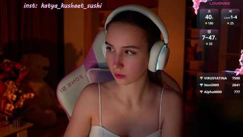 Media: Video of a young woman with fair skin, brown hair, wearing headphones, pink lipstick, and a white tank top, seated in a dimly lit room.