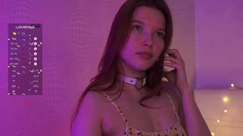 Video of a young Caucasian woman with long brown hair, wearing a white choker and a floral-patterned top, in a dimly-lit room with purple and yellow lighting.