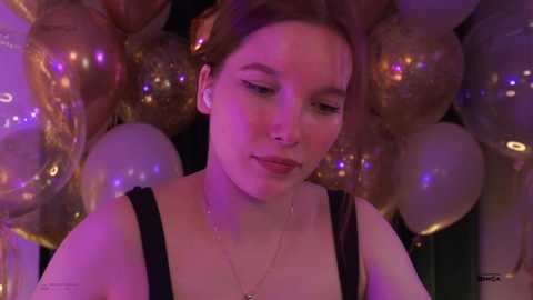 Video of a young woman with fair skin, brown hair, wearing a black tank top, surrounded by metallic balloons in a dimly lit room.