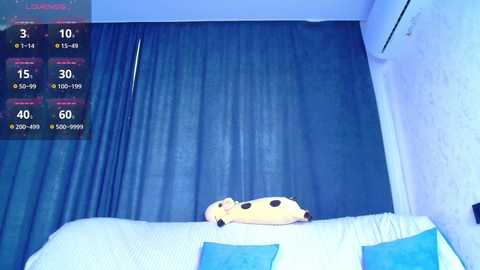 A video of a plush yellow dog toy on a white couch with blue cushions, set against a blue curtain backdrop. A digital display in the top left corner shows a 3D model with stats and options.