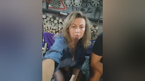 Media: Video of a blonde woman with a serious expression, wearing a blue striped shirt, kneeling in front of a woodpile in a rustic room with wheelbarrow, saw, and metal objects on the wall.