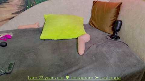 Media: Video of a woman lying on a bed, wearing a green dress, with a life-like male sex doll, a brown pillow, and a black vibrator, in a beige room.