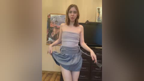 Media: Video of a slender, Caucasian woman with shoulder-length brown hair, wearing a strapless, light grey dress with a high slit, standing in a living room with beige walls, wooden floor, and a dark dresser.