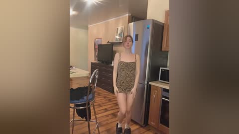 Media: Video of a young Caucasian woman with fair skin and light brown hair in a leopard-print dress, standing in a modern kitchen with wooden floors and stainless steel appliances.