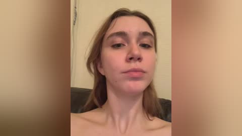 Media: Video of a young Caucasian woman with straight, shoulder-length brown hair, fair skin, and a neutral expression. She appears topless, with a beige wall and dark furniture in the background.