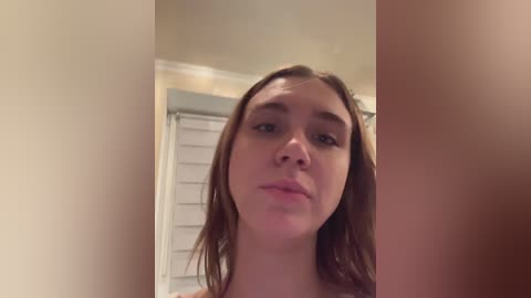 Media: A video of a young Caucasian woman with fair skin, wet brown hair, and light makeup, standing in a dimly lit bathroom with a white window blind and beige walls.