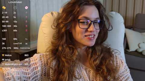 A video of a Caucasian woman with long, wavy brown hair, wearing glasses, a beige knit sweater, and a neutral expression, seated in a beige armchair in a living room.