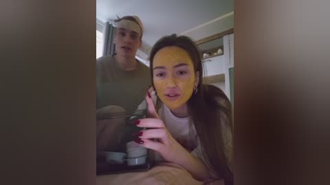 A video captures a young woman with long brown hair, wearing a white top, gesturing with her red-painted nails. Behind her, a young man in a gray hoodie and backwards cap stands, partially obscured by a doorway. The background shows a room with a white ceiling and bookshelves.