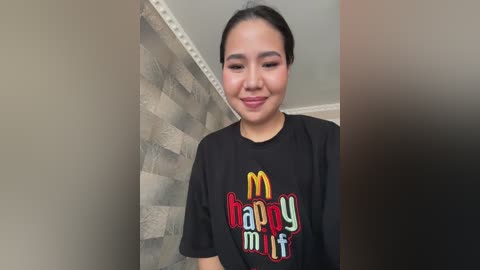 A video of a smiling woman with dark hair and medium skin tone, wearing a black T-shirt with colorful \"McDonald's Happy Meal\" logo. Background features a beige wall and gray brick pattern.
