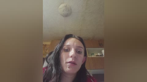 Video of a young woman with long, dark hair, light skin, and a red top, standing in a dimly lit room with a textured ceiling and wooden cabinets.