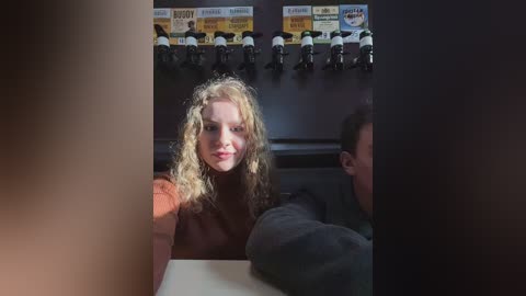 Media: Video of a smiling woman with curly blonde hair, wearing a black top, seated at a table, flanked by two men in dark clothing, in a dimly lit bar with multiple flat-screen TVs displaying sports highlights.