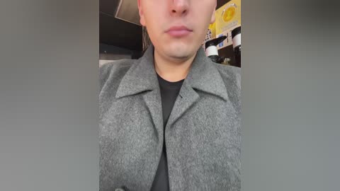 Media: Video of a man in a gray, textured coat with a black shirt underneath, taken indoors with shelves filled with yellow and white boxes in the background.