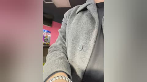 Media: Video of a person wearing a gray, textured coat with a black shirt underneath. The background features a red wall, a shelf with snacks, and a ceiling light.
