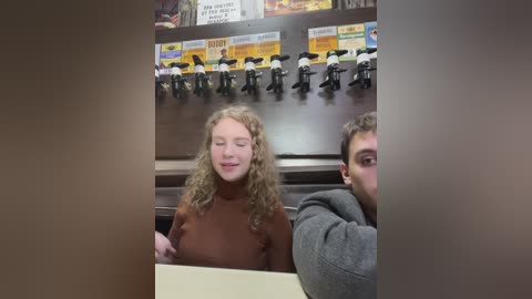 Media: Video of a smiling woman with curly blonde hair and a man with dark hair, both wearing casual attire, sitting in a booth with a menu board above.