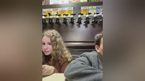 Media: A video of two people in a fast-food restaurant. The woman with curly hair in a brown sweater sits at a table, looking forward. The man in a gray sweater sits beside her, facing away.