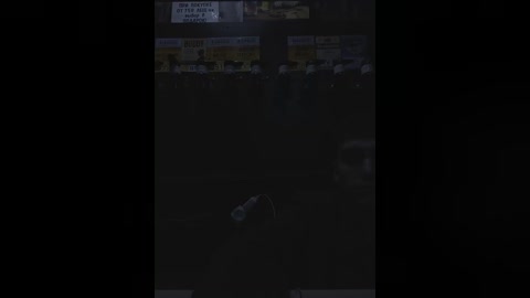 Media: A video of a dark, dimly lit room with a dimly visible figure in a chair, surrounded by rows of people, some obscured by shadows.
