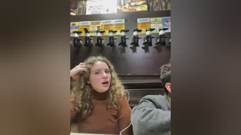 Media: Video of a young girl with curly hair, wearing a brown turtleneck sweater, sitting in a booth. Behind her, a row of black mannequins and a sign displaying prices.