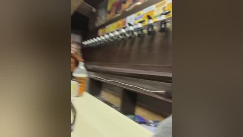 Media: Video of a supermarket beverage aisle with an out-of-focus person in the foreground, holding a white and orange drink bottle. The background showcases a row of soda dispensers with colorful labels.