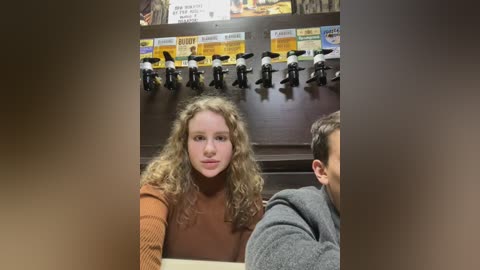 Media: Video of a young girl with curly blonde hair in a brown sweater, sitting in front of a wall with mounted beer taps. A man with short dark hair in a gray hoodie is partially visible in the background.