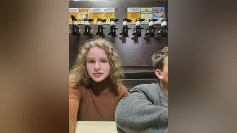 Media: Video of a young woman with curly blonde hair in a brown sweater seated at a table. Background features a wall with mounted toy police officers, and a \"Dunkin' Donuts\" sign.