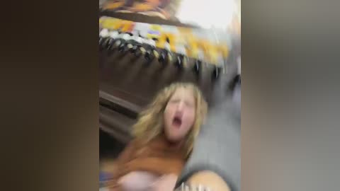 Media: A blurred video shows a blonde woman in an orange sweater, possibly mid-laugh, with a background featuring colorful, hanging items and a table.