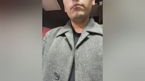 Media: A video of a light-skinned man in a grey wool coat, standing indoors with a red wall in the background. The man's face is partially visible.