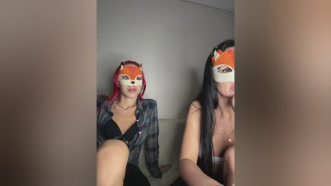 Video: Two women, one with red hair, the other with black hair, both wearing fox masks and plaid shirts, seated on a couch in a dimly lit room.