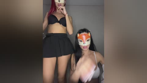 Video of two women: one wearing a black bra and skirt, the other in a red fox mask, topless, with visible breasts, in a dimly lit room.