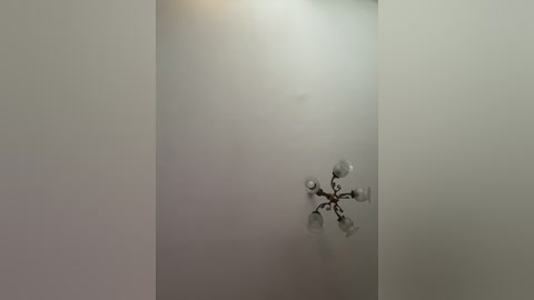 A video of a minimalist, off-white room with a small, ornate, silver chandelier with four bulbs hanging from the ceiling. The chandelier has a vintage, antique design with intricate details. The room's walls are smooth and unadorned, creating a stark contrast with the delicate chandelier.