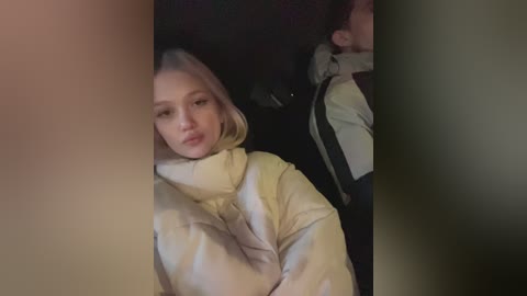 Media: Video of a young woman with fair skin and blonde hair, wearing a yellow hoodie, sitting in a car, kissing someone out of frame.