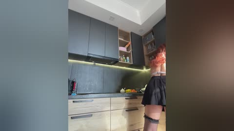 Media: Video of a modern kitchen with a woman in a short black skirt and pink hair, standing near a dark grey cabinet.