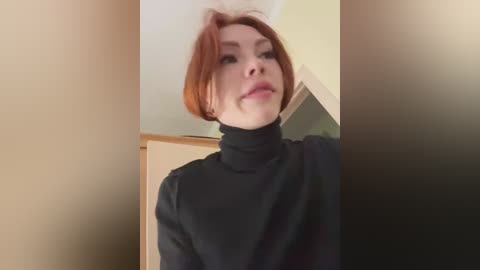 Media: Video of a pale-skinned woman with short, fiery red hair, wearing a black turtleneck sweater, looking slightly to the left, indoors with beige walls and a wooden door in the background.