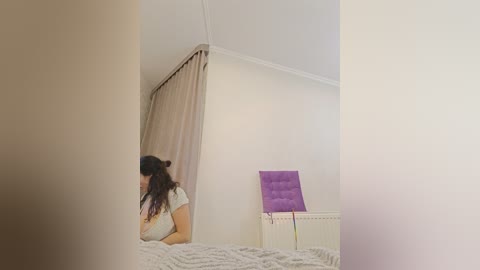 A video of a woman with long dark hair, wearing a white top, sitting on a bed with a light gray blanket, facing a white wall with a purple cushion and a radiator.