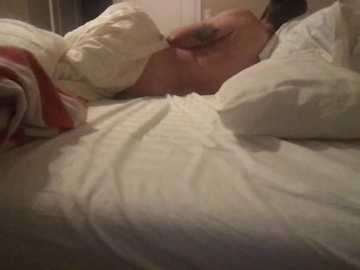Video of a woman lying on a bed, partially covered by white and red blankets. Her left hand rests on her stomach, and her face is not visible. The background is dimly lit.