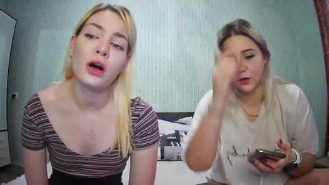 Media: Video of two blonde women, one with a striped top, the other in a white t-shirt, sitting on a bed, looking at a smartphone.