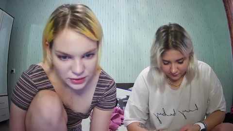 Media: Video of two young women with dyed blonde hair, one in striped top, the other in white t-shirt with \"good and you\" printed, sitting on a bed with light green walls.