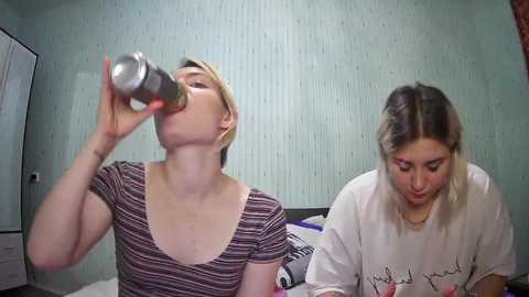 Media: Video of two women, one drinking from a bottle, the other focused on a phone, in a cluttered room with green wallpaper.