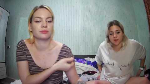 Media: Video of two young women with fair skin, blonde hair, and average build, sitting on a bed in a cluttered room with light blue walls and white furniture.