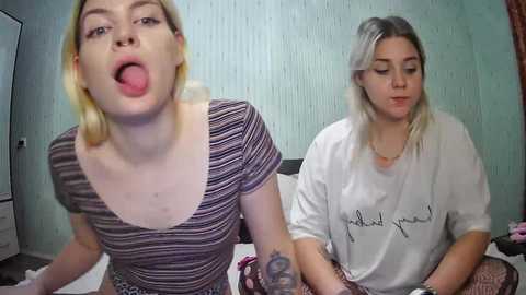 Media: Video of two young women, one blonde with striped shirt, tongue out, and the other with blonde hair in a white shirt, both sitting on a bed.