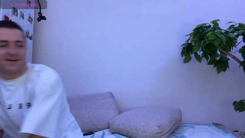Media: Video of a young man in a white shirt, laughing, in a dimly lit room with a green plant, beige pillows, and a white wall.