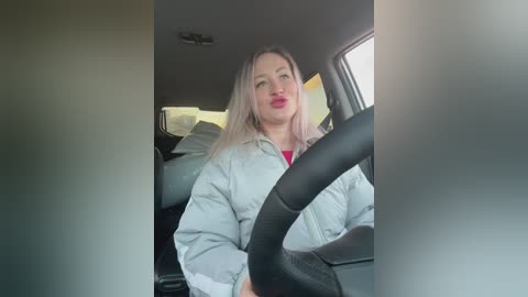 Video of a woman with shoulder-length blonde hair, wearing a gray jacket, driving a car, with the steering wheel in her hands, seen from the back seat.