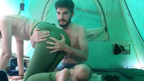 Media: Video of a bearded, shirtless man with a medium build and light skin, sitting inside a green tent. He has a tattoo on his forearm. A woman with light skin and dark hair, wearing a beige tank top and green pants, is bent over him, hands on her thighs.