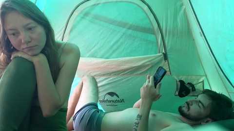 Video of a young Asian woman in a tank top, sitting inside a green tent, looking sad. A shirtless man with a beard, in blue shorts, plays a smartphone inside the tent.