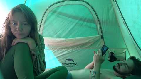 A video shows a young woman with long hair and a man in a tent; she's inside, looking sad, while he's outside, holding a phone.