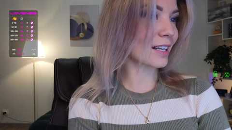 Media: Video of a smiling woman with light brown hair in a green and white striped sweater, seated in a modern living room with a lit lamp, a black chair, and abstract art on the wall.