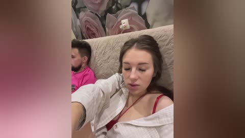 Media: A video of a young woman with light skin and long brown hair in a white robe, sitting on a beige couch. A man with a beard and pink shirt sits beside her. A floral wallpaper background is visible.